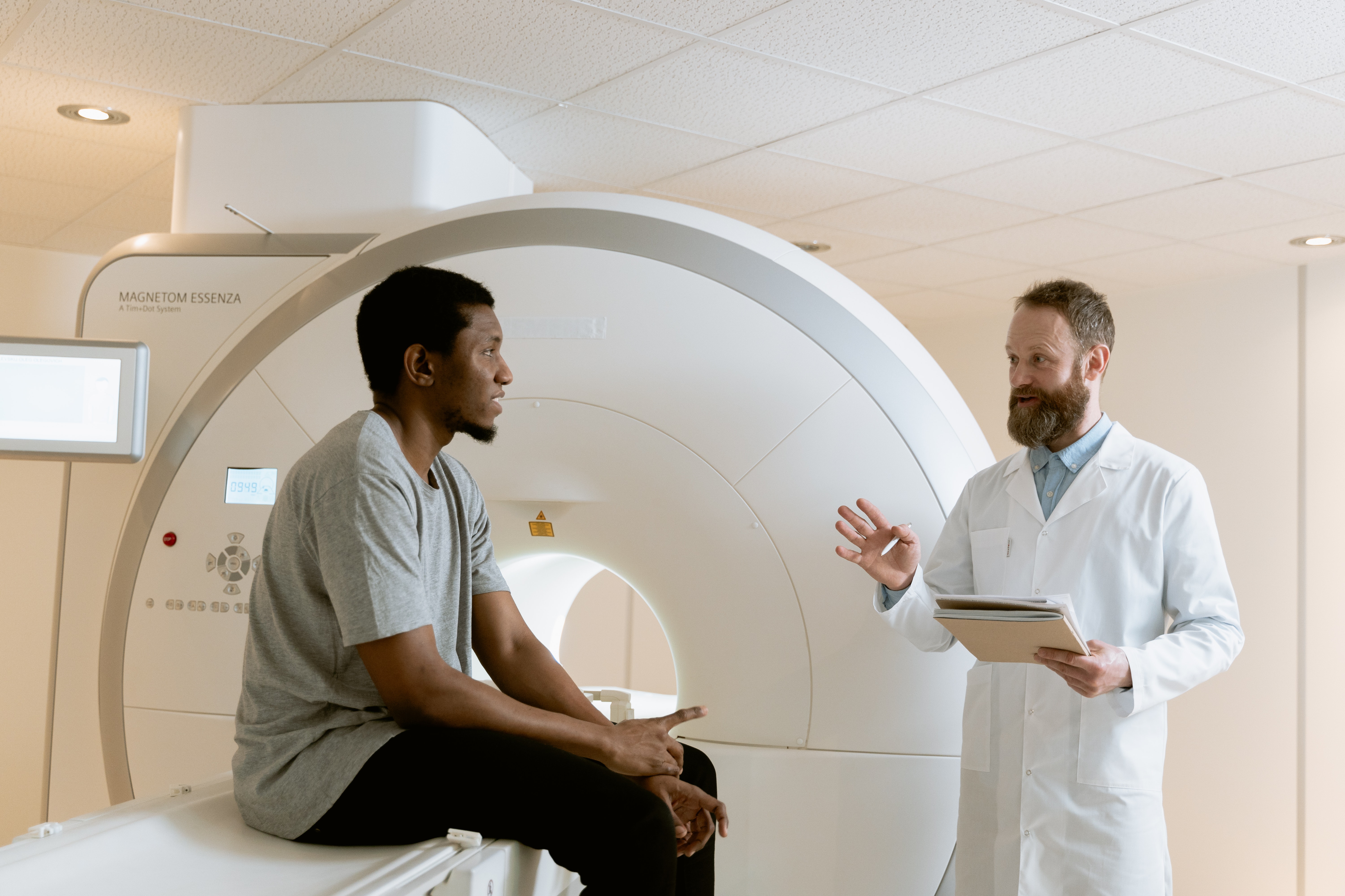 A man sitting on the bed of an MRI scanner is talking to another man in a medical blouse