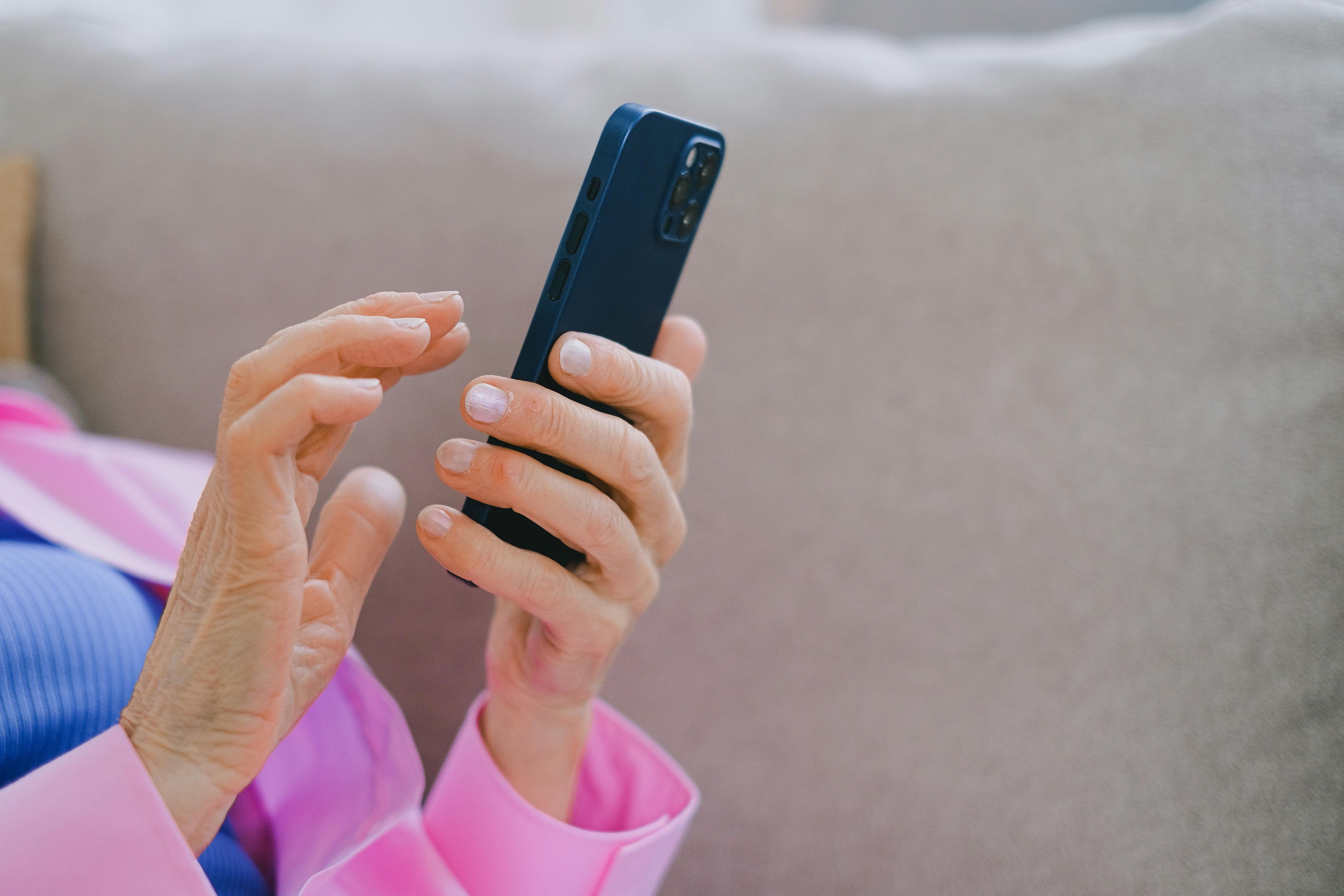 A close up of an elderly woman's hands holding and typing on a smartphone. The woman is wearing a bright pink shirt.
