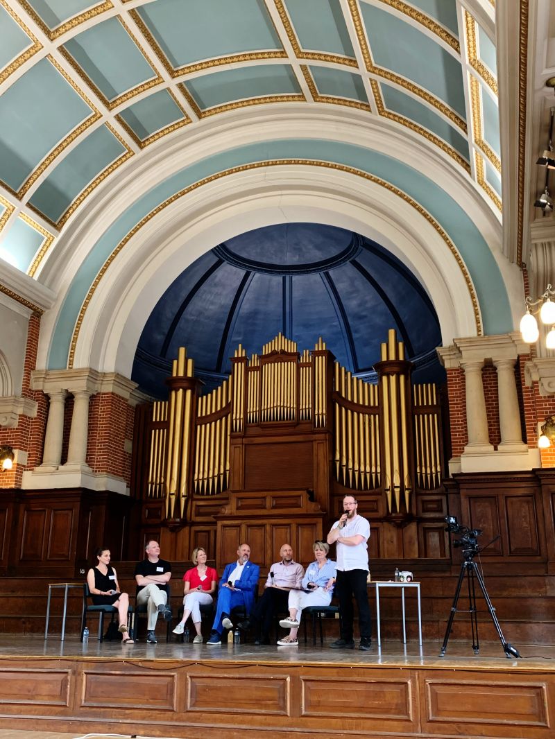 The photo shows six people sat on chairs and one standing, speaking in a microphone, on a stage in the University of Reading's Great Hall