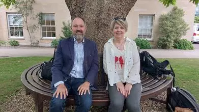 Prof Andrew Rice and BBC Radio presenter Fiona Talkington sitting on a bench under a tree, facing the camera