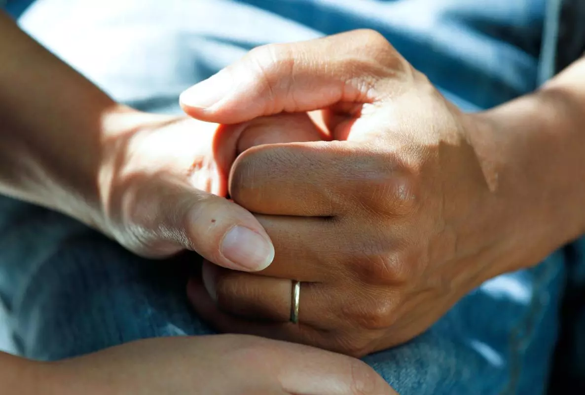 Two hands gently clasp each other, conveying support and compassion. One hand wears a simple ring.