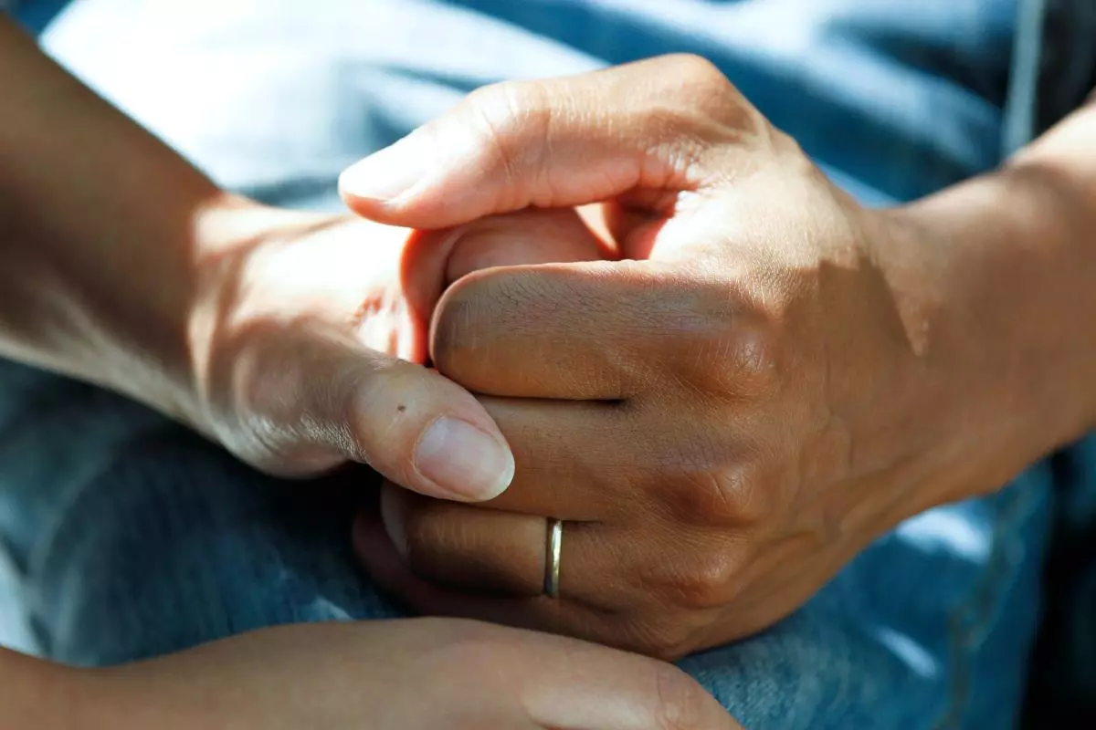 Two hands gently clasp each other, conveying support and compassion. One hand wears a simple ring.